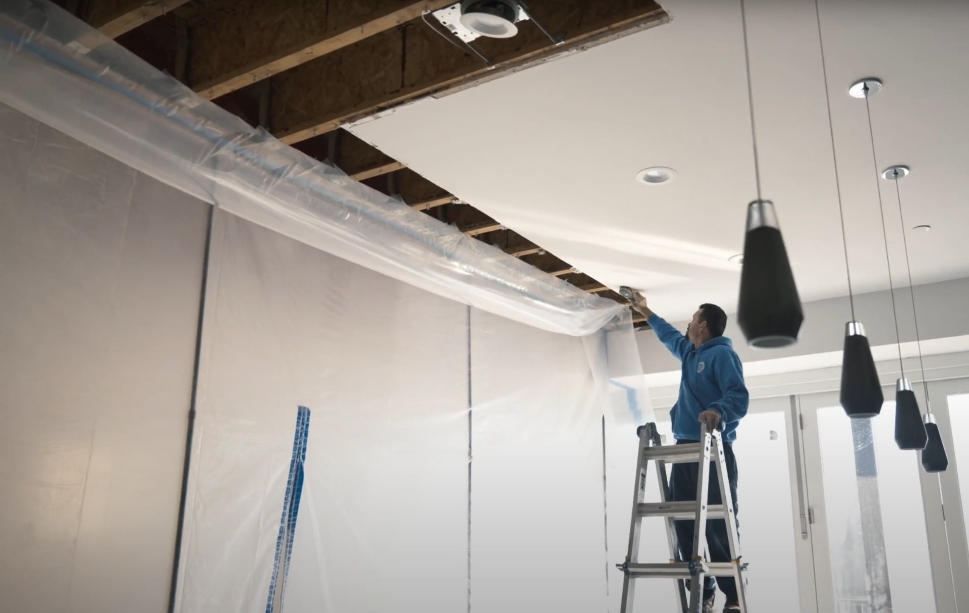 Professional water damage restoration technician working on ceiling repair with plastic sheeting and ladder in commercial space
